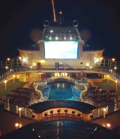 A luxury cruise ship deck featuring a pool surrounded by lounge chairs and warm ambient lighting. A large screen overlooks the area, showing an image against a dark night sky.