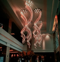 A luxurious interior space featuring an extravagant chandelier with swirling, illuminated designs hanging from a high ceiling. The area is warmly lit, with elegant furnishings and large vases containing decorative plants. Soft lighting and modern decor create a sophisticated atmosphere.