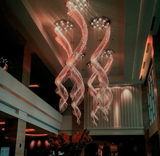 A luxurious interior space featuring an extravagant chandelier with swirling, illuminated designs hanging from a high ceiling. The area is warmly lit, with elegant furnishings and large vases containing decorative plants. Soft lighting and modern decor create a sophisticated atmosphere.