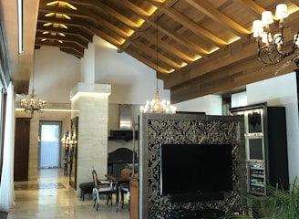 The image displays a luxurious interior with a high wooden vaulted ceiling featuring modern lighting. There is an ornate partition with a large flat-screen TV, surrounded by stylish decor. The room includes elegant chandeliers, a dining table with chairs, and plant decor, creating a sophisticated and cozy atmosphere.