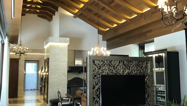 The image displays a luxurious interior with a high wooden vaulted ceiling featuring modern lighting. There is an ornate partition with a large flat-screen TV, surrounded by stylish decor. The room includes elegant chandeliers, a dining table with chairs, and plant decor, creating a sophisticated and cozy atmosphere.