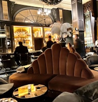 A luxurious lounge with elegant, plush seating and a sophisticated bar. The interior features dark wood paneling, ornate lighting fixtures, and a stunning backdrop of illuminated bottles on shelves. The atmosphere is warm and inviting, with various patrons sitting at the bar and others moving around the space. Large decorative plumes and an opulent chandelier add to the grandeur of the room.