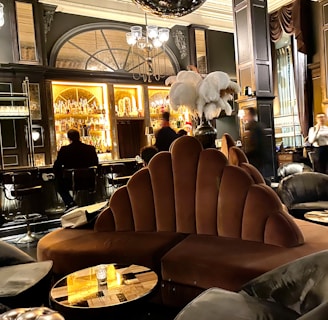 A luxurious lounge with elegant, plush seating and a sophisticated bar. The interior features dark wood paneling, ornate lighting fixtures, and a stunning backdrop of illuminated bottles on shelves. The atmosphere is warm and inviting, with various patrons sitting at the bar and others moving around the space. Large decorative plumes and an opulent chandelier add to the grandeur of the room.