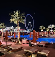 A nighttime scene of a luxurious outdoor pool area with orange umbrellas and loungers, illuminated by palm trees and accent lights. In the background, a large Ferris wheel glows with blue and white lights, enhancing the vibrant cityscape. The setting suggests a relaxing and upscale atmosphere.