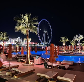 A nighttime scene of a luxurious outdoor pool area with orange umbrellas and loungers, illuminated by palm trees and accent lights. In the background, a large Ferris wheel glows with blue and white lights, enhancing the vibrant cityscape. The setting suggests a relaxing and upscale atmosphere.