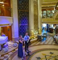 A luxurious interior space with a grand staircase and ornate decor. A small musical ensemble, including a violinist and a singer, performs near a white grand piano. Several people are seated, watching the performance, while others casually walk by. The setting appears elegant and relaxed.