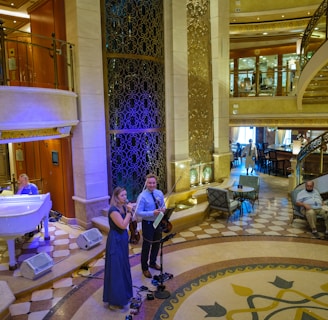 A luxurious interior space with a grand staircase and ornate decor. A small musical ensemble, including a violinist and a singer, performs near a white grand piano. Several people are seated, watching the performance, while others casually walk by. The setting appears elegant and relaxed.