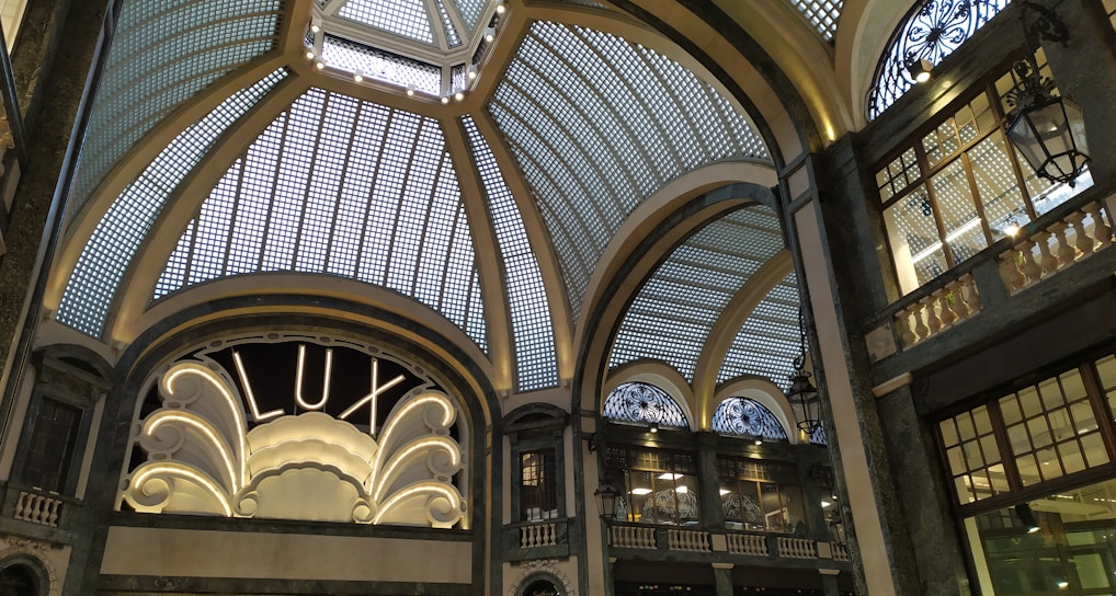A grand, ornate interior with an elaborate glass ceiling composed of geometric panels, adorned with decorative metalwork. An elegant sign reading 'LUX' is illuminated, framed by intricate architectural details. Dim lighting casts soft shadows, enhancing the sophisticated ambiance.