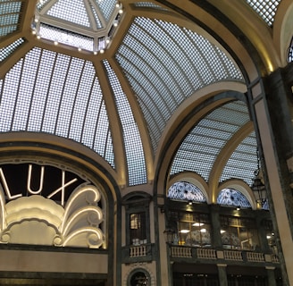 A grand, ornate interior with an elaborate glass ceiling composed of geometric panels, adorned with decorative metalwork. An elegant sign reading 'LUX' is illuminated, framed by intricate architectural details. Dim lighting casts soft shadows, enhancing the sophisticated ambiance.