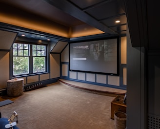 A cozy home theater room features a large projection screen displaying a movie selection interface. The room has dark wood paneling with soft, warm lighting highlighting the ceiling and walls. A large window is situated on one side with an outdoor view partially obscured by trees. There is a curved platform below the screen and a comfortable seating area, including a chevron-patterned ottoman. The room has a neutral color palette with beige, dark gray, and soft brown tones.