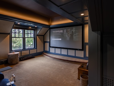A cozy home theater room features a large projection screen displaying a movie selection interface. The room has dark wood paneling with soft, warm lighting highlighting the ceiling and walls. A large window is situated on one side with an outdoor view partially obscured by trees. There is a curved platform below the screen and a comfortable seating area, including a chevron-patterned ottoman. The room has a neutral color palette with beige, dark gray, and soft brown tones.
