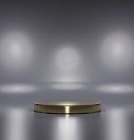 A metallic platform is centered in a minimalistic setting with a reflective surface. Soft, diffused lighting creates circular patterns on the background wall, enhancing the focus on the central stage. The ambiance is sleek and modern, with a hint of elegance.