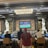 Several people are seated at a luxurious bar, with a large TV screen showing a sports game. The bar is elegantly decorated with ornate details and sculptures. The ceiling has a large, rectangular, crystal chandelier.