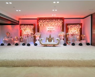 An elegant and luxurious stage setup featuring a lavishly decorated sofa in the center, surrounded by ornate floral arrangements. The backdrop is adorned with cascading flowers and soft, ambient lighting that emits a warm glow. The overall decor is rich with intricate details, creating an opulent and festive atmosphere.