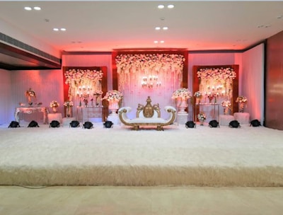 An elegant and luxurious stage setup featuring a lavishly decorated sofa in the center, surrounded by ornate floral arrangements. The backdrop is adorned with cascading flowers and soft, ambient lighting that emits a warm glow. The overall decor is rich with intricate details, creating an opulent and festive atmosphere.