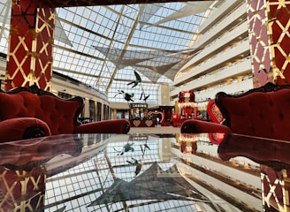 A luxurious interior space featuring plush red armchairs and sofas against a backdrop of a large glass ceiling, allowing natural light to illuminate the area. The furniture is placed around a reflective table surface, enhancing the opulent and grand ambiance. The decor includes elaborate golden patterns on red pillars.