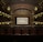 A grand, ornate theater with a large screen at the center, surrounded by elegantly designed architectural elements. The interior features intricate patterns and warm lighting, with multiple levels of seating and decorative balconies. The atmosphere suggests a historic or classic design theme.