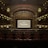 A grand, ornate theater with a large screen at the center, surrounded by elegantly designed architectural elements. The interior features intricate patterns and warm lighting, with multiple levels of seating and decorative balconies. The atmosphere suggests a historic or classic design theme.