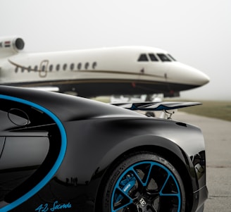A luxury sports car with sleek black bodywork and blue accents is prominently featured in the foreground, showcasing its high-performance wheels. In the background, a private jet is parked on an overcast day, highlighting a scene of opulence and sophistication.