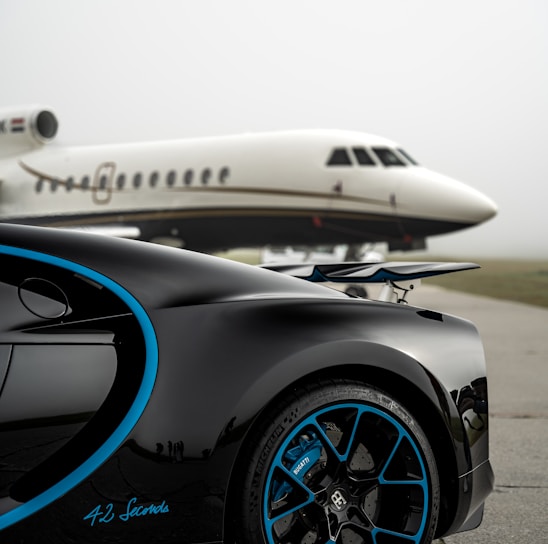 A luxury sports car with sleek black bodywork and blue accents is prominently featured in the foreground, showcasing its high-performance wheels. In the background, a private jet is parked on an overcast day, highlighting a scene of opulence and sophistication.