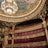 An opulent theater interior featuring grand architectural details with intricate gold ornamentation. A massive chandelier hangs from the ornate ceiling, which displays colorful artwork. The seating area is plush, with rows of luxurious red seats.