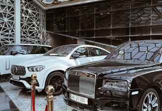 A luxurious indoor setting with a geometric glass ceiling and reflective marble floor. Three high-end luxury cars, including a black and white color scheme, are prominently displayed. A red velvet rope separates the cars from the viewer, suggesting exclusivity.
