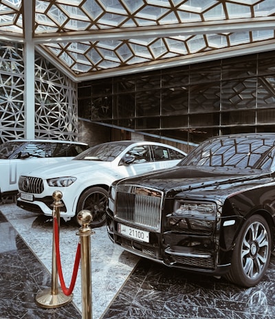 A luxurious indoor setting with a geometric glass ceiling and reflective marble floor. Three high-end luxury cars, including a black and white color scheme, are prominently displayed. A red velvet rope separates the cars from the viewer, suggesting exclusivity.